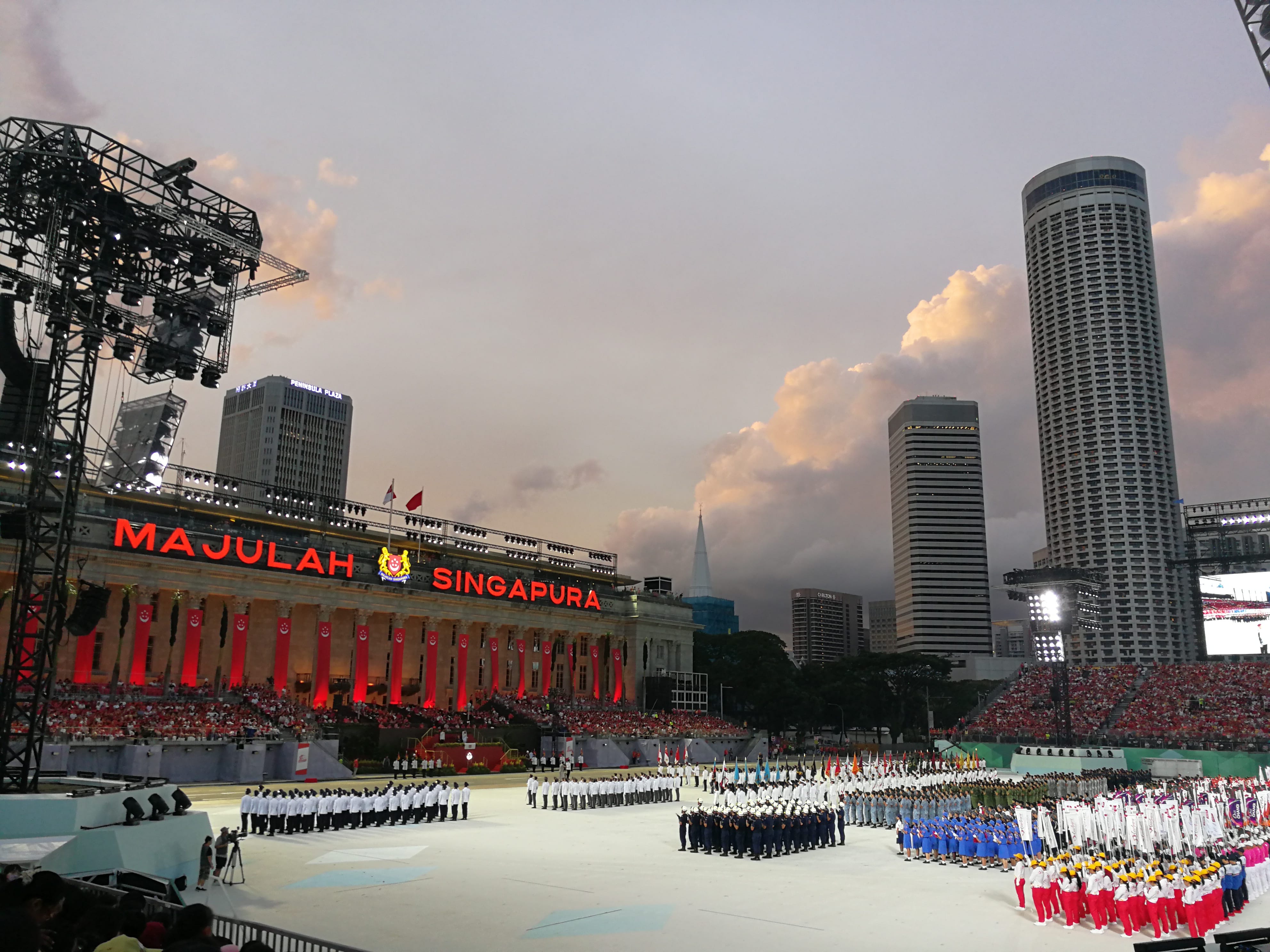 Large parade at Singapore's Padang with "Majulah Singapura" lit on the City Hall facade at dusk.